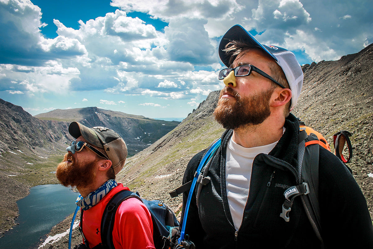 Two rugged outdoor men hiking the mountains wearing Noz-up blue and yellow sunscreen, odorless skunk, and bear repellent on their noses.